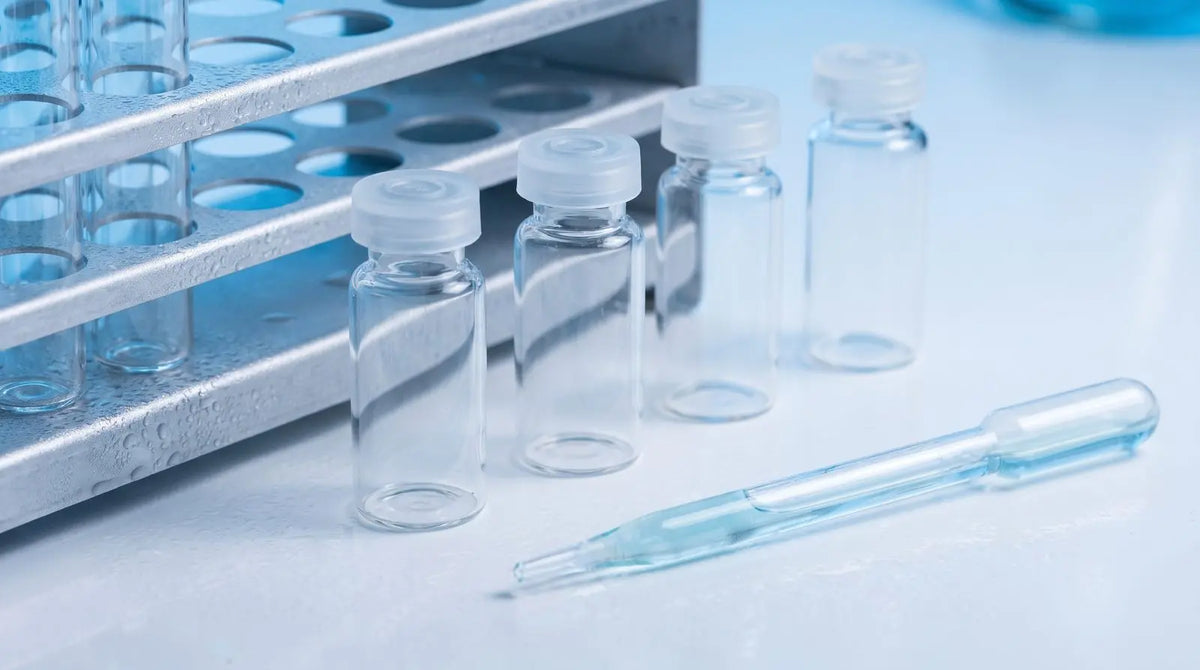 Clear glass laboratory vials beside a glass pipette and a frosted metal rack in cool blue tones, representing cold-storage handling and reconstitution of research peptides.